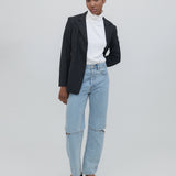 A person stands against a plain background wearing a black blazer, white turtleneck, and the light blue Cowgirl Jeans, which feature knee slits. They complete the look with black shoes, have a neutral expression, and keep one hand in their pocket.