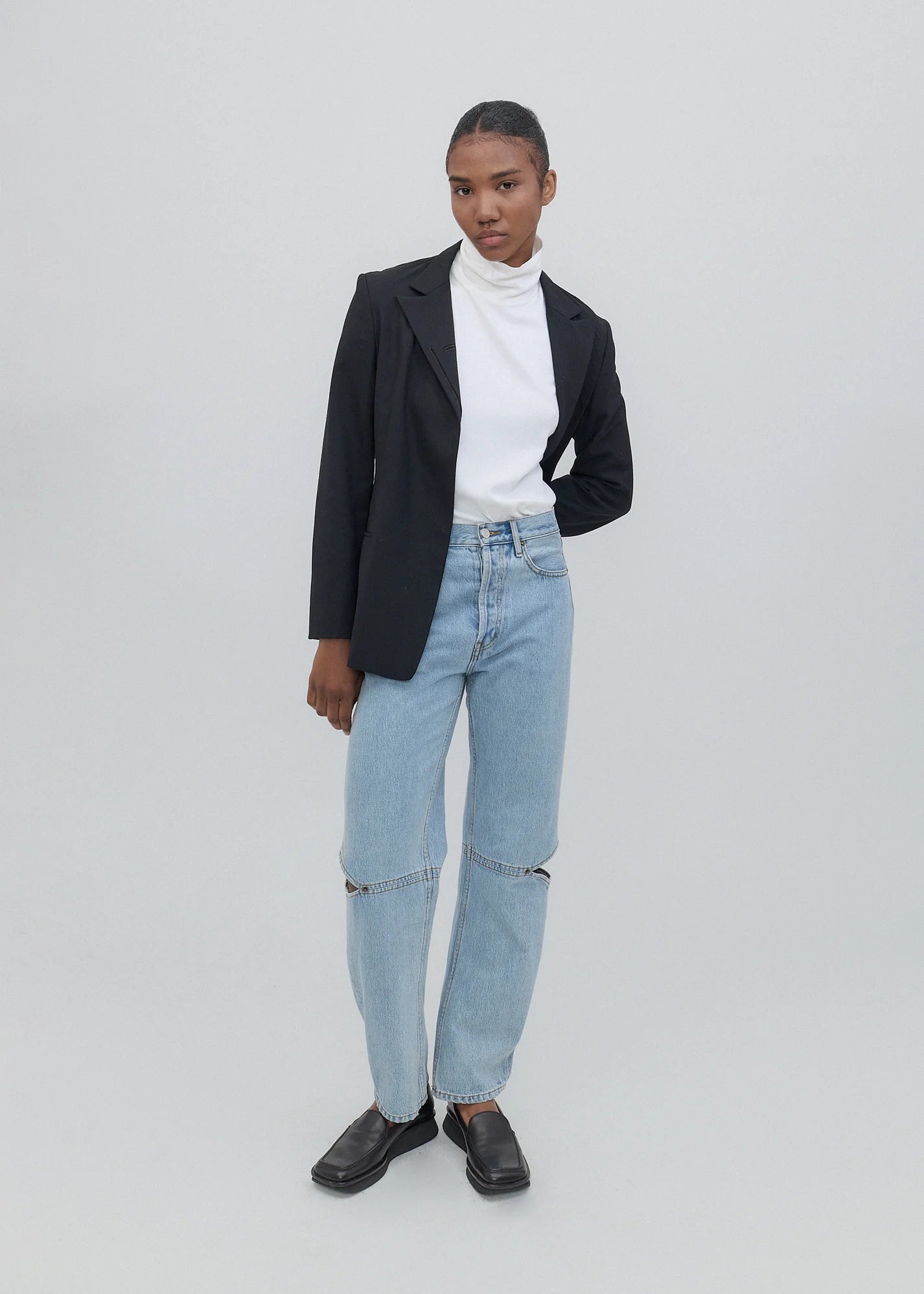 A person stands against a plain background wearing a black blazer, white turtleneck, and the light blue Cowgirl Jeans, which feature knee slits. They complete the look with black shoes, have a neutral expression, and keep one hand in their pocket.