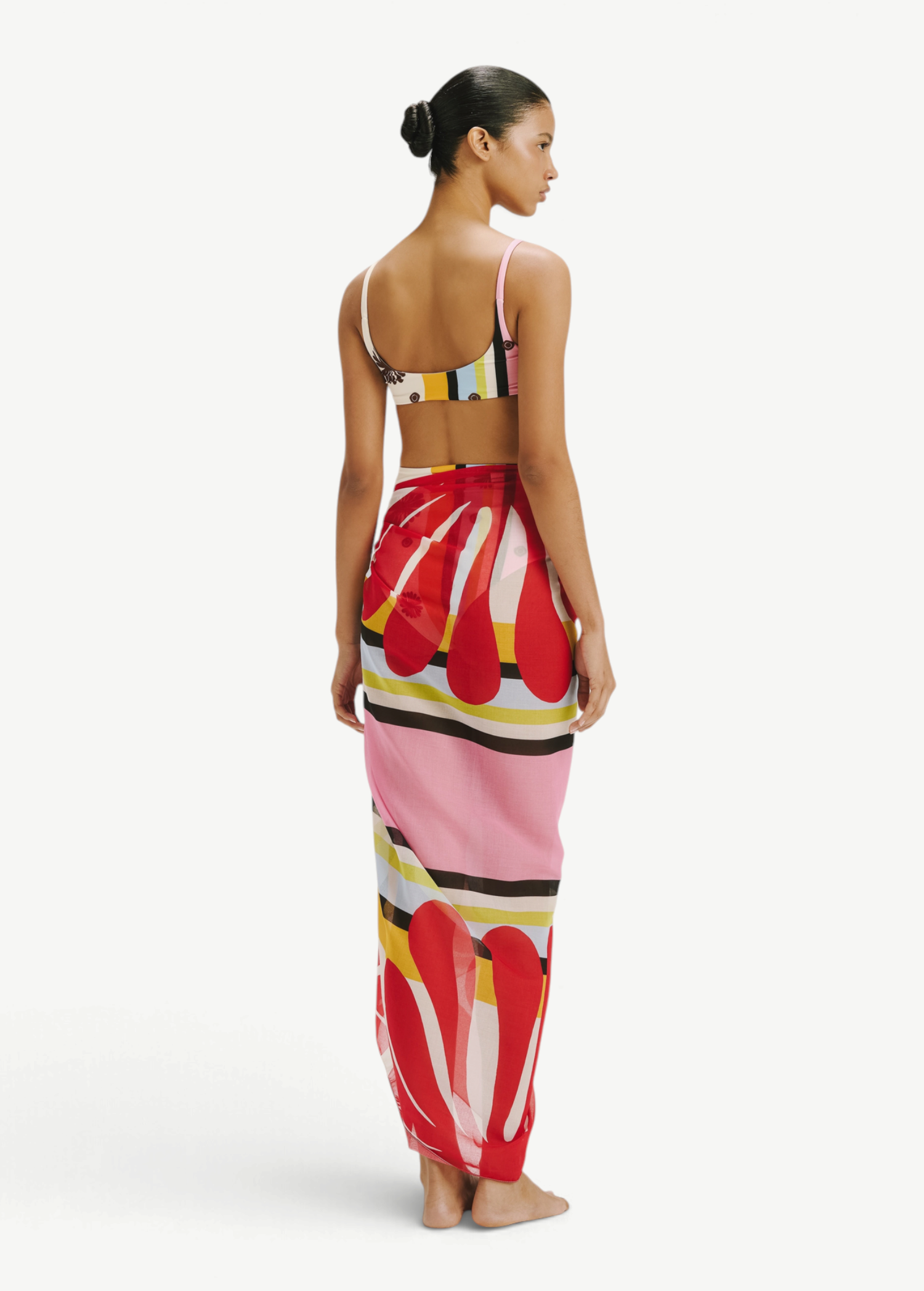 Model wearing a KABE SHOP INC Luana Sarong featuring a vibrant red, pink, and yellow abstract print over swimwear.