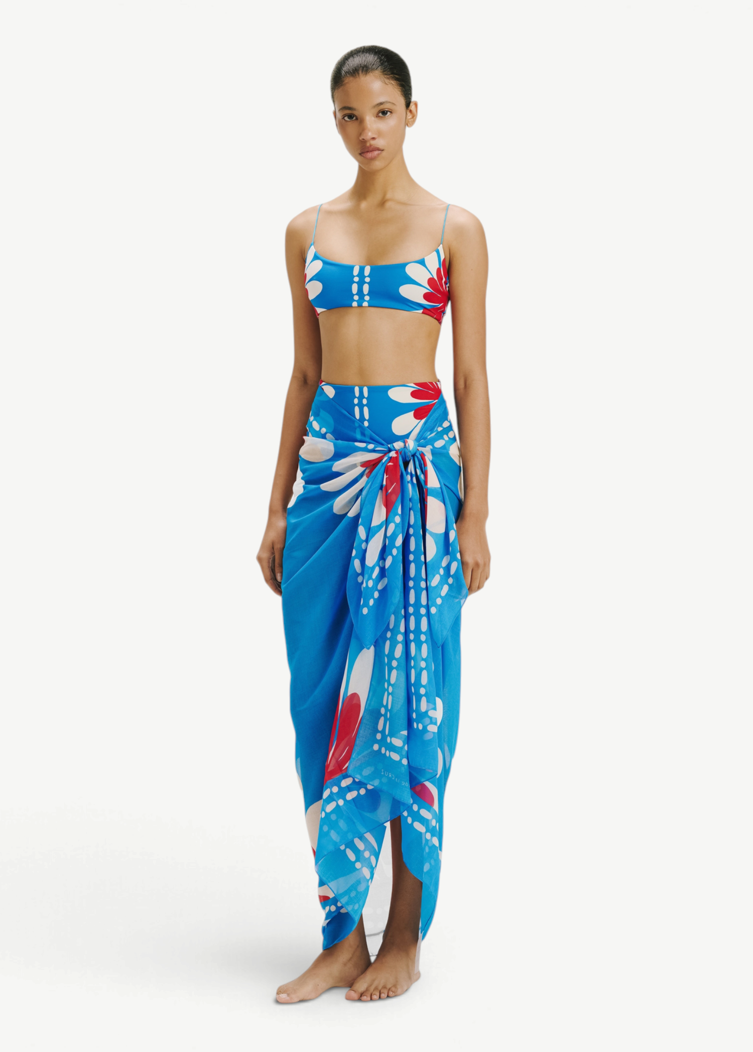 Model wearing a KABE SHOP INC Luana Sarong in blue with a vibrant red and white floral print over matching swimwear.