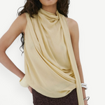 A model wears the Menorca Top, a pale yellow sleeveless piece with a draped cowl neckline, paired with a dark brown lace skirt in an asymmetric silhouette.