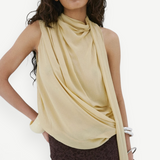A model wears the Menorca Top, a pale yellow sleeveless piece with a draped cowl neckline, paired with a dark brown lace skirt in an asymmetric silhouette.