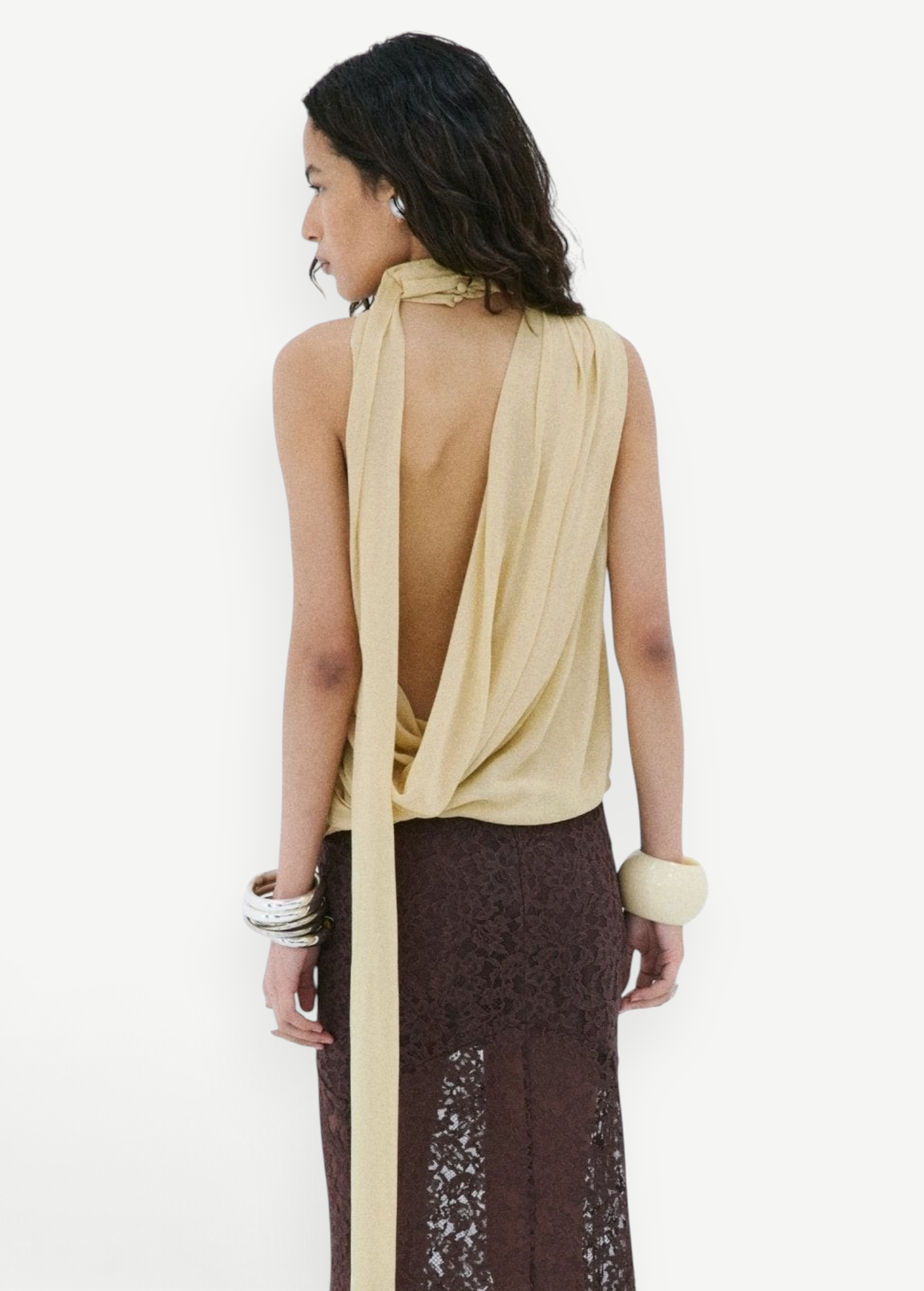 A person wears a pale yellow Menorca Top in viscose georgette with a draped open back and a long brown lace skirt.