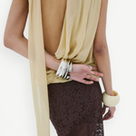 A person wearing a yellow Menorca top in viscose georgette with a backless, draped silhouette, paired with a dark brown lace skirt and stacked silver and cream-colored bracelets.