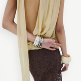 A person wearing a yellow Menorca top in viscose georgette with a backless, draped silhouette, paired with a dark brown lace skirt and stacked silver and cream-colored bracelets.
