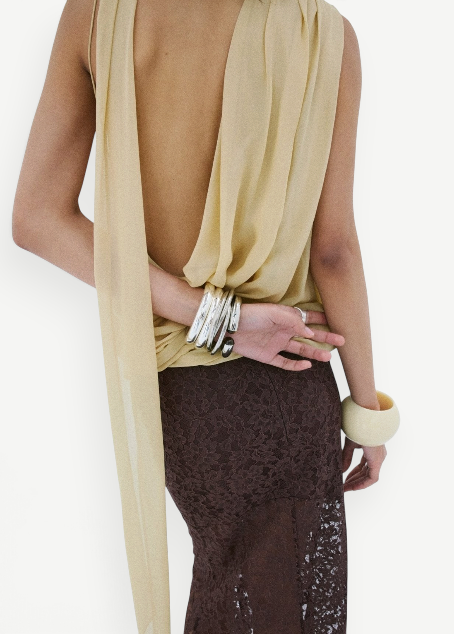 A person wearing a yellow Menorca top in viscose georgette with a backless, draped silhouette, paired with a dark brown lace skirt and stacked silver and cream-colored bracelets.