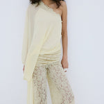 A woman stands against a plain white background, looking directly at the camera, wearing the Elia Top by De La Vali—a cream, off-shoulder design in fluid georgette with elegant draping and a single long sleeve—paired with high-waisted, wide-leg sheer lace pants.