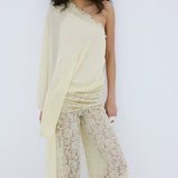 A woman stands against a plain white background, looking directly at the camera, wearing the Elia Top by De La Vali—a cream, off-shoulder design in fluid georgette with elegant draping and a single long sleeve—paired with high-waisted, wide-leg sheer lace pants.