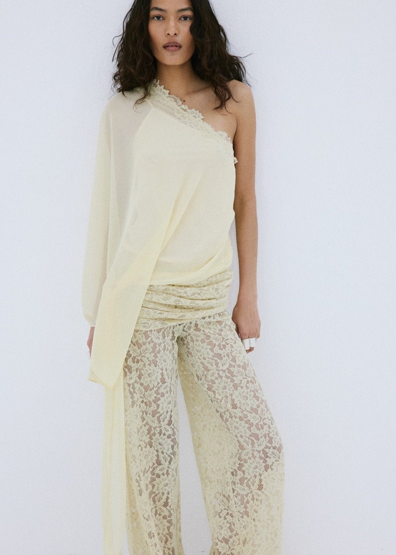 A woman stands against a plain white background, looking directly at the camera, wearing the Elia Top by De La Vali—a cream, off-shoulder design in fluid georgette with elegant draping and a single long sleeve—paired with high-waisted, wide-leg sheer lace pants.