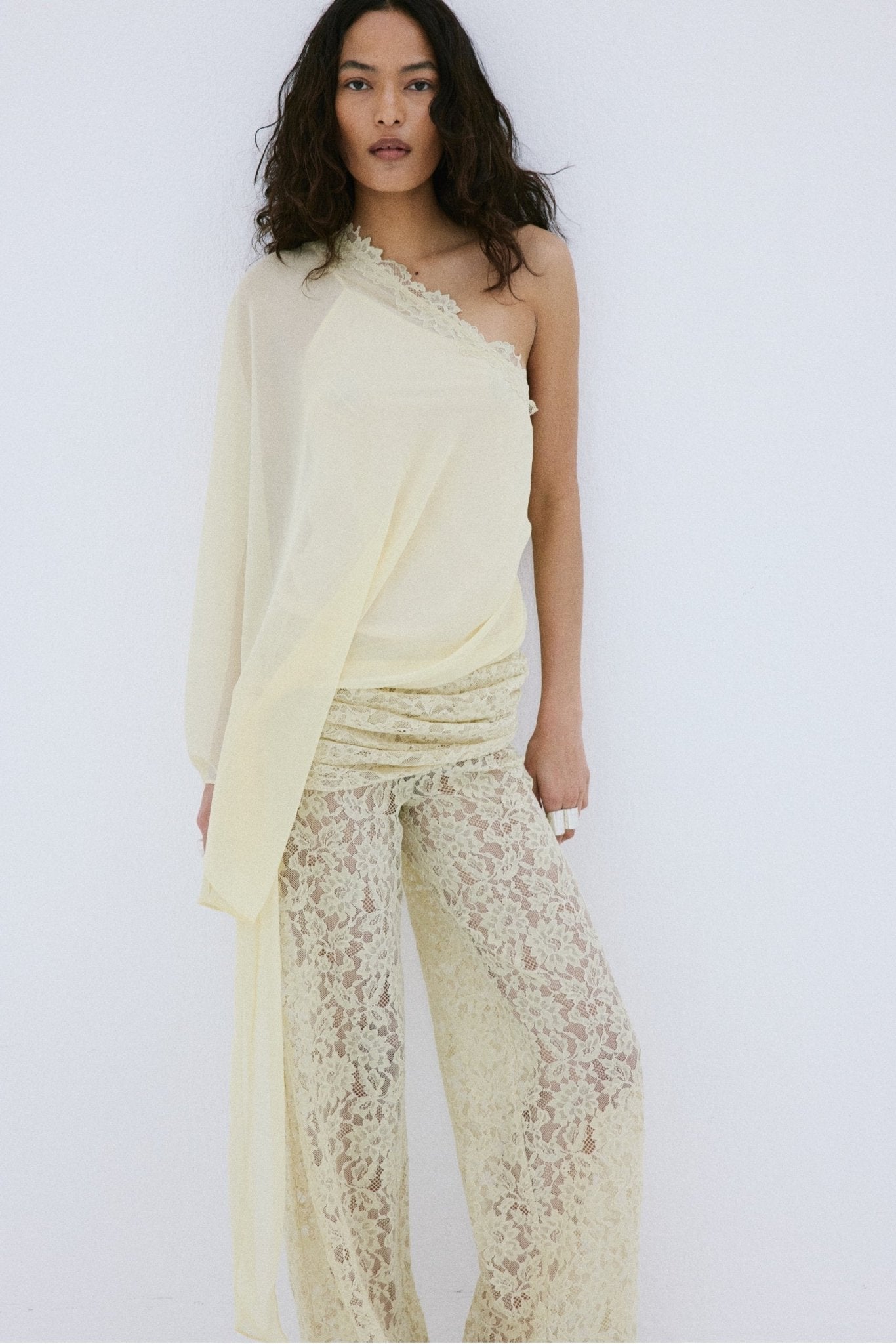 A woman stands against a plain white background, looking directly at the camera, wearing the Elia Top by De La Vali—a cream, off-shoulder design in fluid georgette with elegant draping and a single long sleeve—paired with high-waisted, wide-leg sheer lace pants.