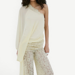 A woman stands against a plain white background, looking directly at the camera, wearing the Elia Top by De La Vali—a cream, off-shoulder design in fluid georgette with elegant draping and a single long sleeve—paired with high-waisted, wide-leg sheer lace pants.