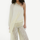 A woman stands against a plain white background, looking directly at the camera, wearing the Elia Top by De La Vali—a cream, off-shoulder design in fluid georgette with elegant draping and a single long sleeve—paired with high-waisted, wide-leg sheer lace pants.