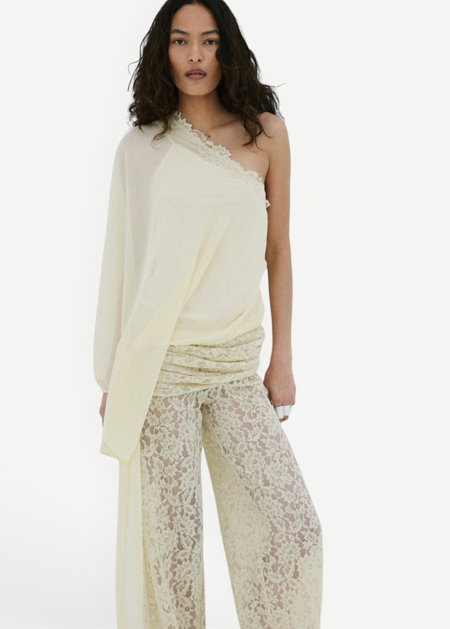 A woman stands against a plain white background, looking directly at the camera, wearing the Elia Top by De La Vali—a cream, off-shoulder design in fluid georgette with elegant draping and a single long sleeve—paired with high-waisted, wide-leg sheer lace pants.