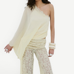 A woman wearing the Elia Top—a fluid georgette, one-shoulder design—paired with cream lace pants, a beaded headpiece, and chunky bangles stands against a plain white background.