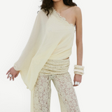 A woman wearing the Elia Top—a fluid georgette, one-shoulder design—paired with cream lace pants, a beaded headpiece, and chunky bangles stands against a plain white background.