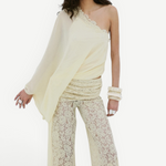 A model wears pale yellow Rocio Trouser lace trousers in a vintage-inspired draped silhouette, paired with a matching one-shoulder top, lace headpiece, and stacked bangles.