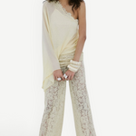 A woman stands against a plain white background, wearing the Elia Top in cream from De La Vali—a fluid georgette one-shoulder style—paired with sheer lace pants. She has long wavy hair, accessorizes with bracelets, and completes the look with black sandals.