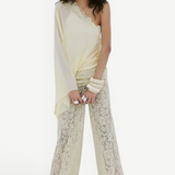 A woman stands against a plain white background, wearing the Elia Top in cream from De La Vali—a fluid georgette one-shoulder style—paired with sheer lace pants. She has long wavy hair, accessorizes with bracelets, and completes the look with black sandals.
