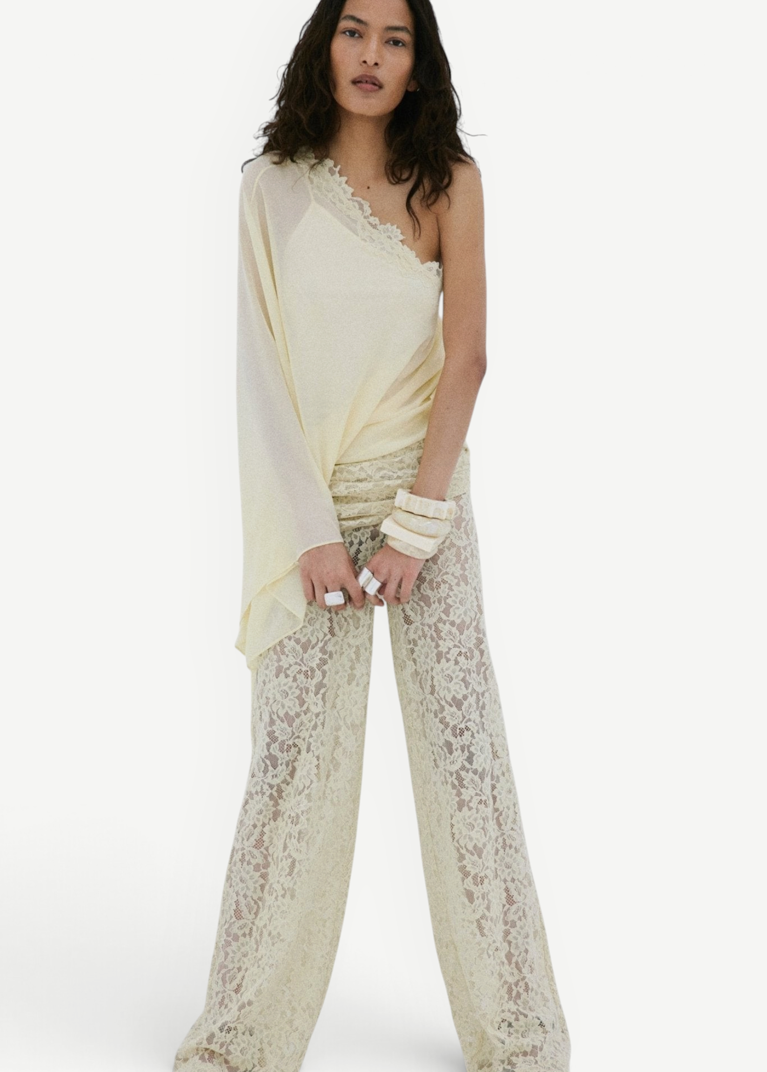 A woman stands against a plain white background, wearing the Elia Top in cream from De La Vali—a fluid georgette one-shoulder style—paired with sheer lace pants. She has long wavy hair, accessorizes with bracelets, and completes the look with black sandals.