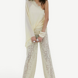 A person wearing a vintage-inspired, pale yellow, one-shoulder lace top and matching Rocio Trouser with a draped, wide-leg silhouette.
