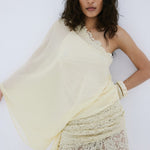 A woman with medium-length wavy hair and a neutral expression poses against a white wall, wearing De La Vali’s Elia Top—a one-shoulder, flowing piece in light yellow georgette—paired with matching lace pants.