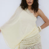 A woman with medium-length wavy hair and a neutral expression poses against a white wall, wearing De La Vali’s Elia Top—a one-shoulder, flowing piece in light yellow georgette—paired with matching lace pants.