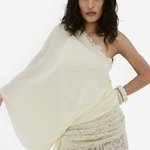 A woman with medium-length wavy hair and a neutral expression poses against a white wall, wearing De La Vali’s Elia Top—a one-shoulder, flowing piece in light yellow georgette—paired with matching lace pants.