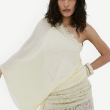 A woman with medium-length wavy hair and a neutral expression poses against a white wall, wearing De La Vali’s Elia Top—a one-shoulder, flowing piece in light yellow georgette—paired with matching lace pants.