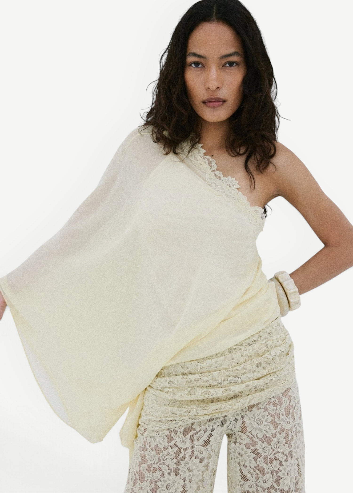 A woman with medium-length wavy hair and a neutral expression poses against a white wall, wearing De La Vali’s Elia Top—a one-shoulder, flowing piece in light yellow georgette—paired with matching lace pants.