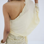 A person stands with their back turned, modeling the off-shoulder Elia Top in lacy, off-white fabric by De La Vali. Their left hand is raised near their face, hair styled in loose waves, and they wear matching lace pants. The backdrop is plain white.