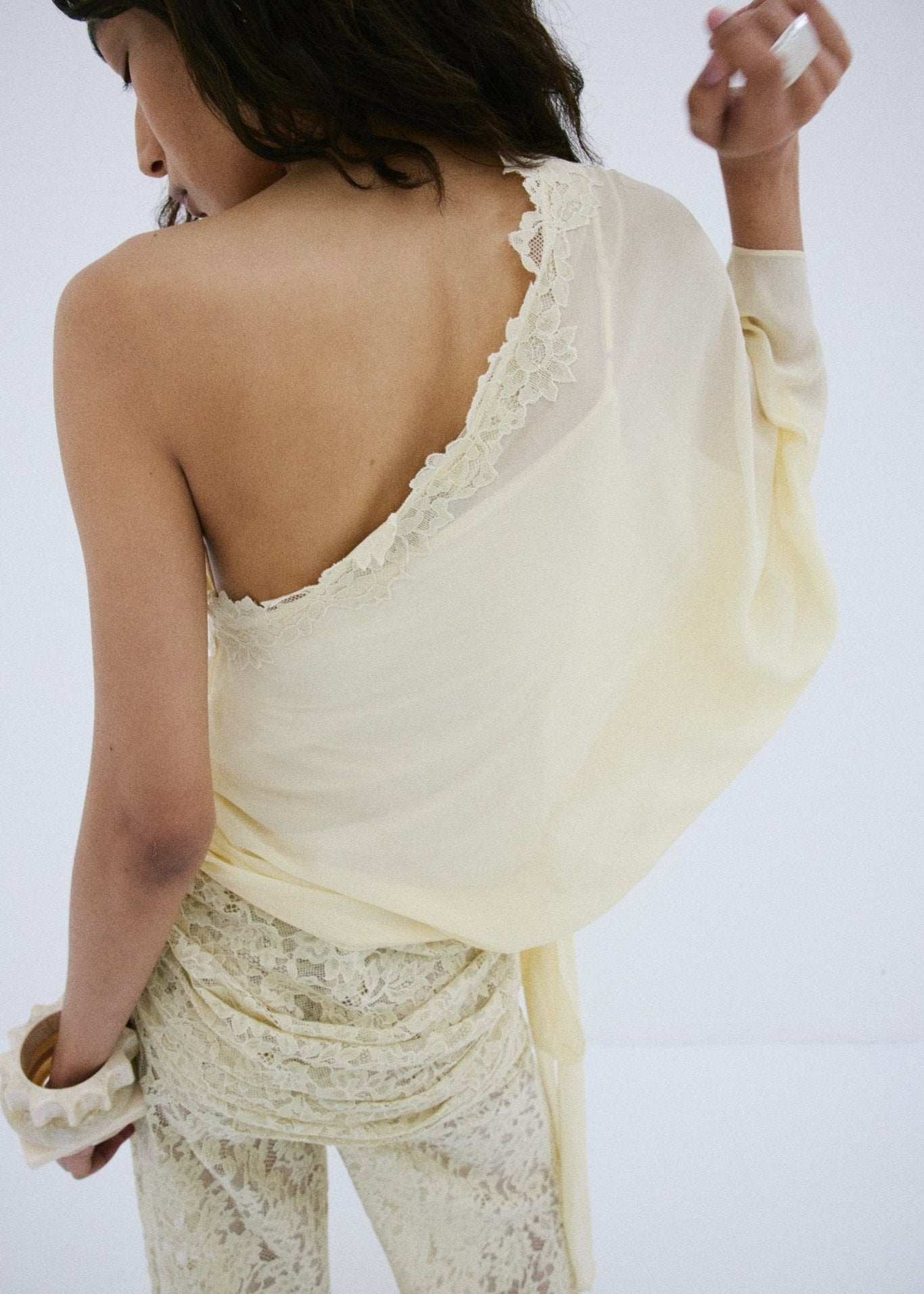 A person stands with their back turned, modeling the off-shoulder Elia Top in lacy, off-white fabric by De La Vali. Their left hand is raised near their face, hair styled in loose waves, and they wear matching lace pants. The backdrop is plain white.