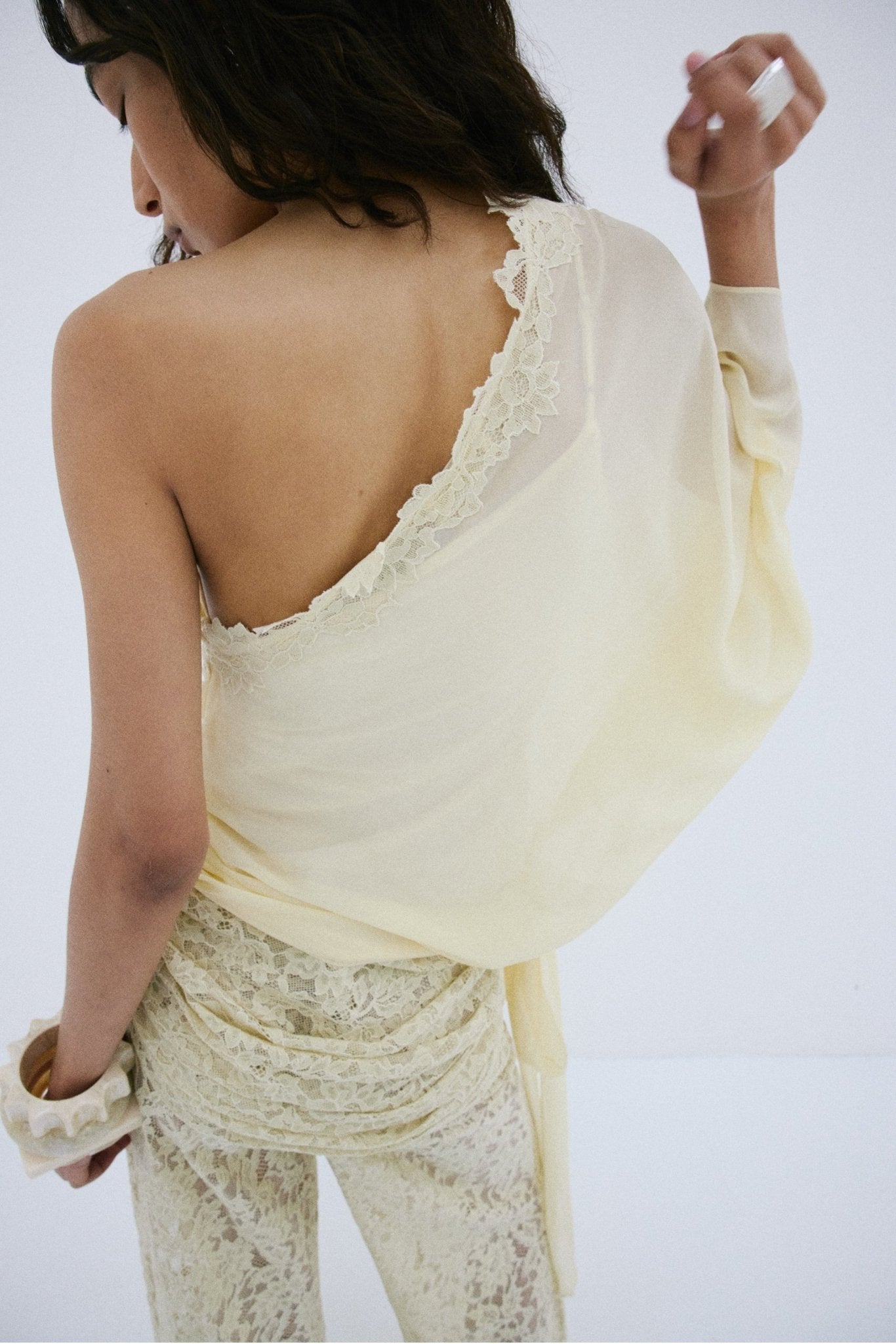 A person stands with their back turned, modeling the off-shoulder Elia Top in lacy, off-white fabric by De La Vali. Their left hand is raised near their face, hair styled in loose waves, and they wear matching lace pants. The backdrop is plain white.