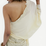 A person stands with their back turned, modeling the off-shoulder Elia Top in lacy, off-white fabric by De La Vali. Their left hand is raised near their face, hair styled in loose waves, and they wear matching lace pants. The backdrop is plain white.