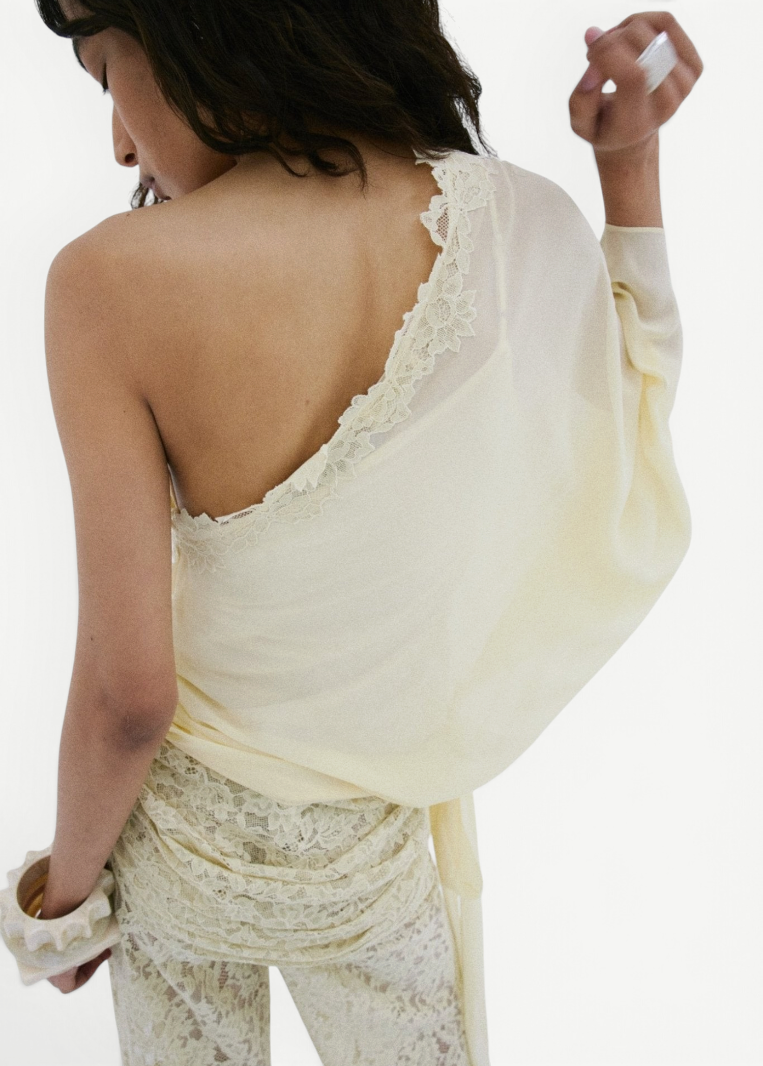 A person stands with their back turned, modeling the off-shoulder Elia Top in lacy, off-white fabric by De La Vali. Their left hand is raised near their face, hair styled in loose waves, and they wear matching lace pants. The backdrop is plain white.