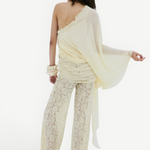 A person seen from behind, wearing a cream-colored, one-shoulder sheer top with a draped silhouette and matching Rocio Trouser lace wide-leg trousers.