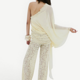 A person seen from behind, wearing a cream-colored, one-shoulder sheer top with a draped silhouette and matching Rocio Trouser lace wide-leg trousers.