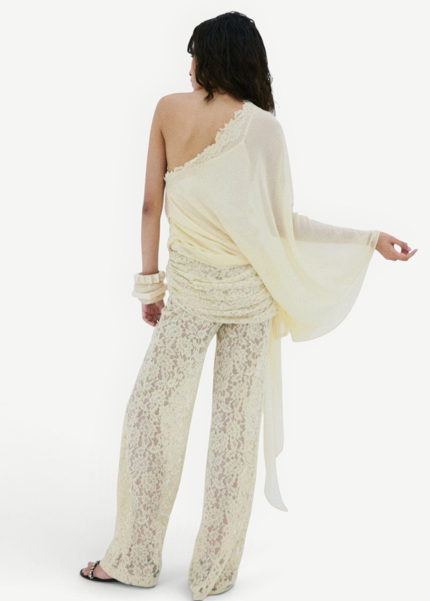 A person seen from behind, wearing a cream-colored, one-shoulder sheer top with a draped silhouette and matching Rocio Trouser lace wide-leg trousers.
