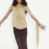 A person wears a pale yellow Menorca top in a sleeveless, draped viscose georgette with a long scarf detail and a floor-length brown lace skirt.