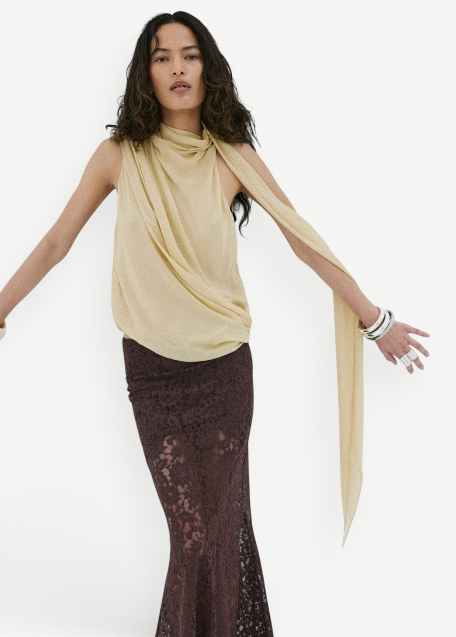 A person wears a pale yellow Menorca top in a sleeveless, draped viscose georgette with a long scarf detail and a floor-length brown lace skirt.