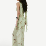 A person seen from the side wearing the Diosa Skirt, a light green, asymmetric hem maxi skirt with a floral pattern and a matching sleeveless top with long, draped shoulder ties.