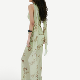 A person seen from the side wearing the Diosa Skirt, a light green, asymmetric hem maxi skirt with a floral pattern and a matching sleeveless top with long, draped shoulder ties.