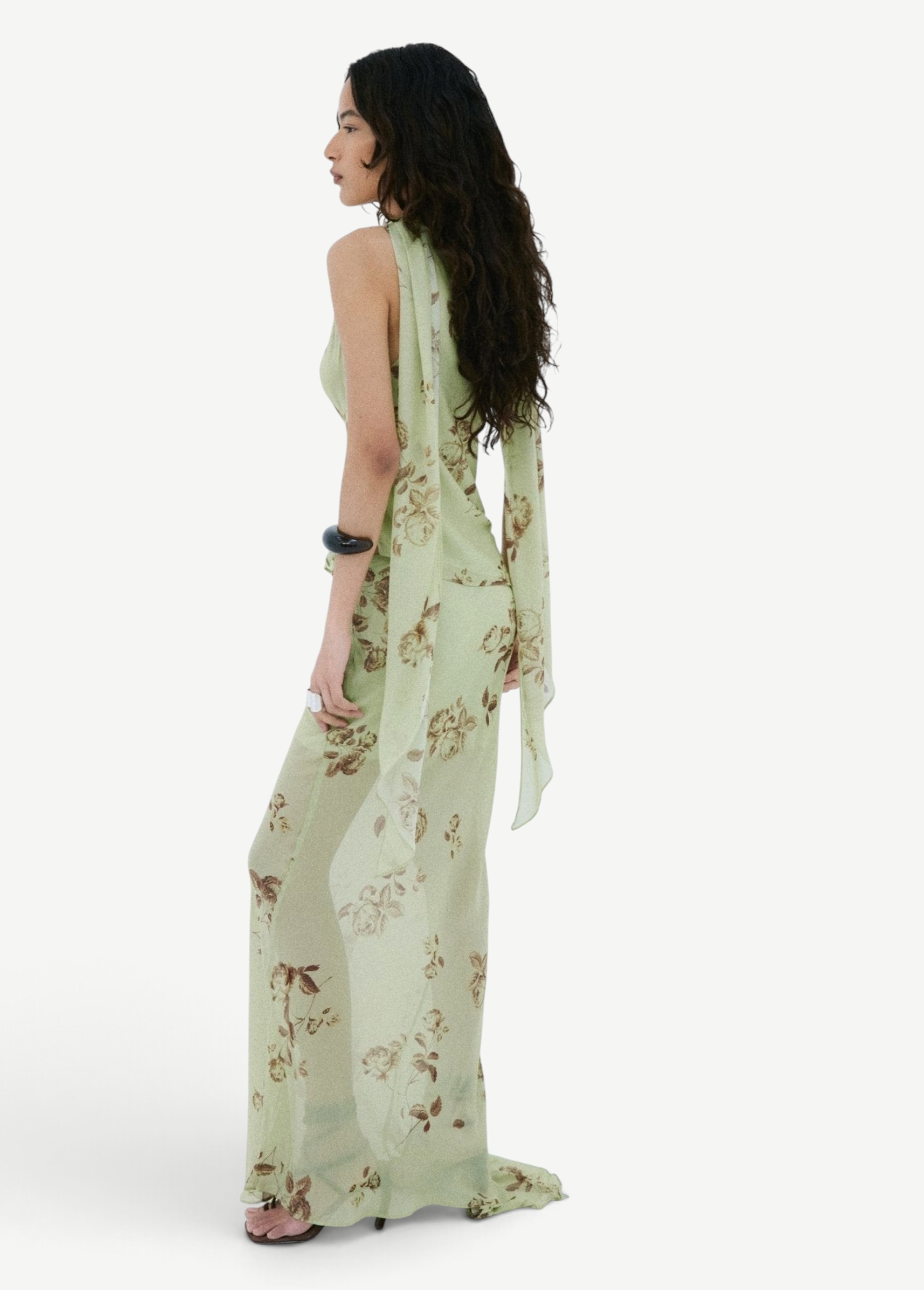 A person seen from the side wearing the Diosa Skirt, a light green, asymmetric hem maxi skirt with a floral pattern and a matching sleeveless top with long, draped shoulder ties.
