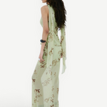A person seen from the side wearing the Diosa Skirt, a light green, asymmetric hem maxi skirt with a floral pattern and a matching sleeveless top with long, draped shoulder ties.