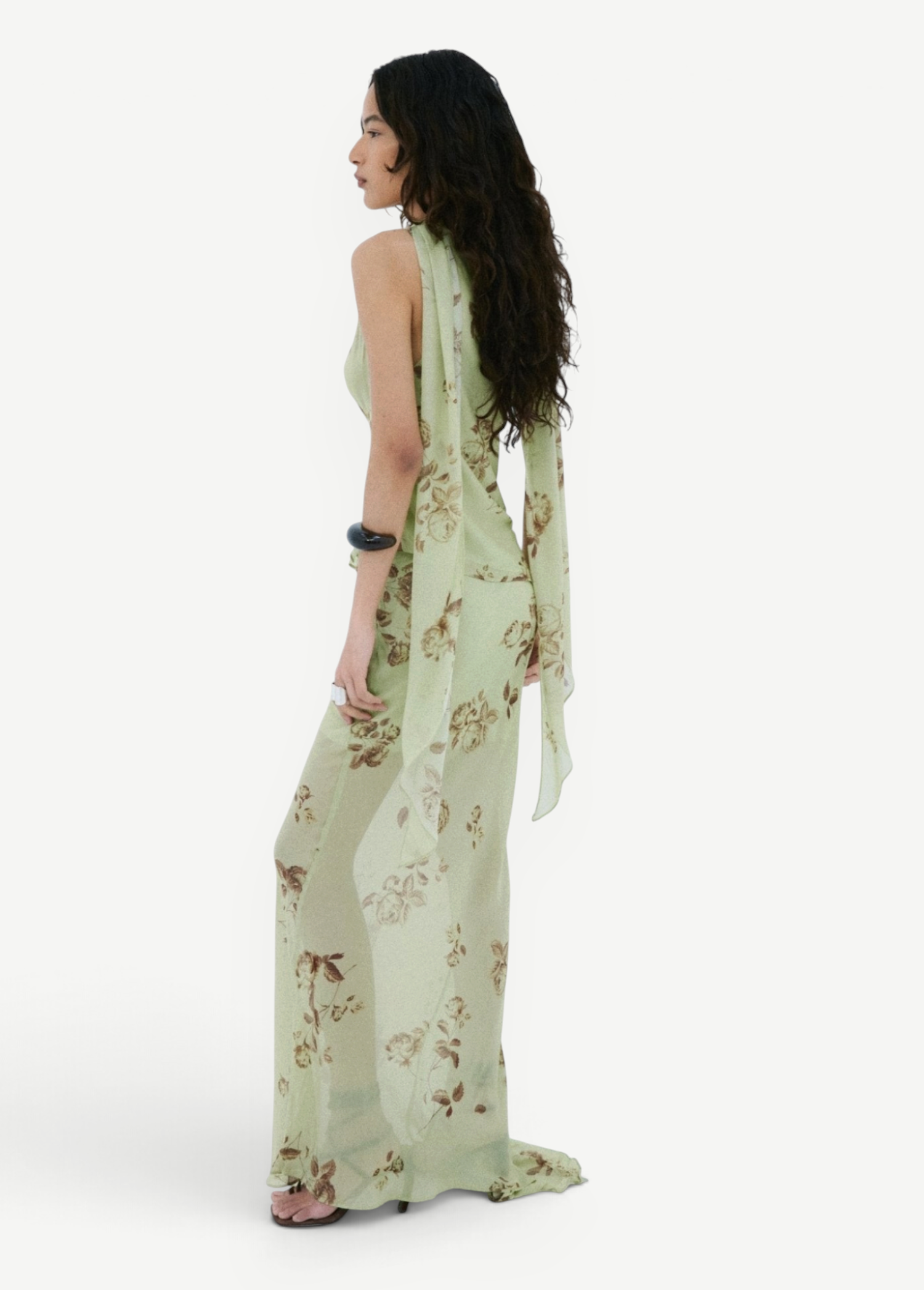 A person seen from the side wearing the Diosa Skirt, a light green, asymmetric hem maxi skirt with a floral pattern and a matching sleeveless top with long, draped shoulder ties.