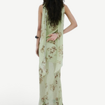 A person stands against a white wall wearing a floor-length, light green floral Diosa Skirt, featuring an asymmetric hem and a draped panel down the back.
