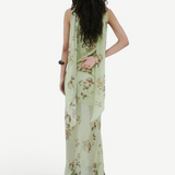A person stands against a white wall wearing a floor-length, light green floral Diosa Skirt, featuring an asymmetric hem and a draped panel down the back.