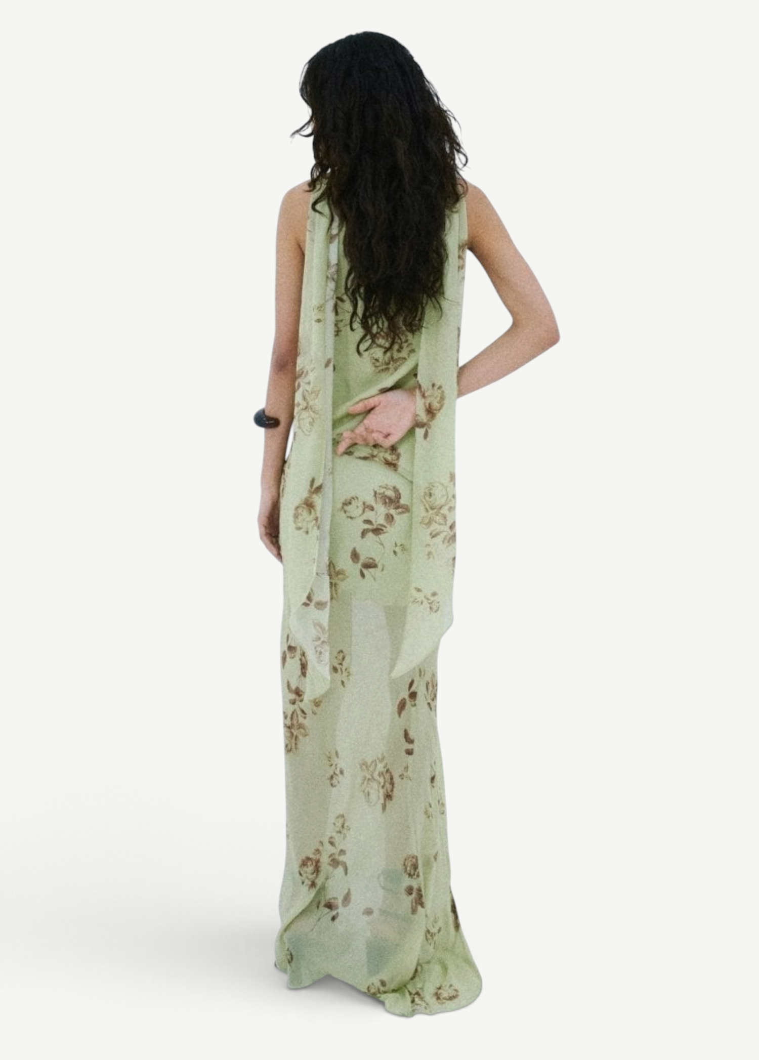 A person stands against a white wall wearing a floor-length, light green floral Diosa Skirt, featuring an asymmetric hem and a draped panel down the back.