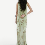 A person stands against a white wall wearing a floor-length, light green floral Diosa Skirt, featuring an asymmetric hem and a draped panel down the back.