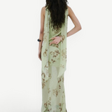 A person stands against a white wall wearing a floor-length, light green floral Diosa Skirt, featuring an asymmetric hem and a draped panel down the back.