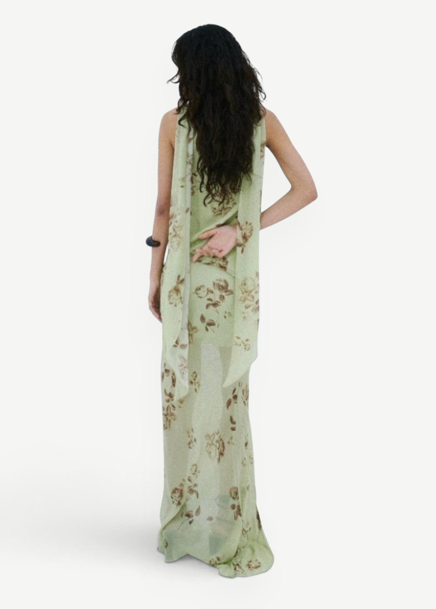 A person stands against a white wall wearing a floor-length, light green floral Diosa Skirt, featuring an asymmetric hem and a draped panel down the back.